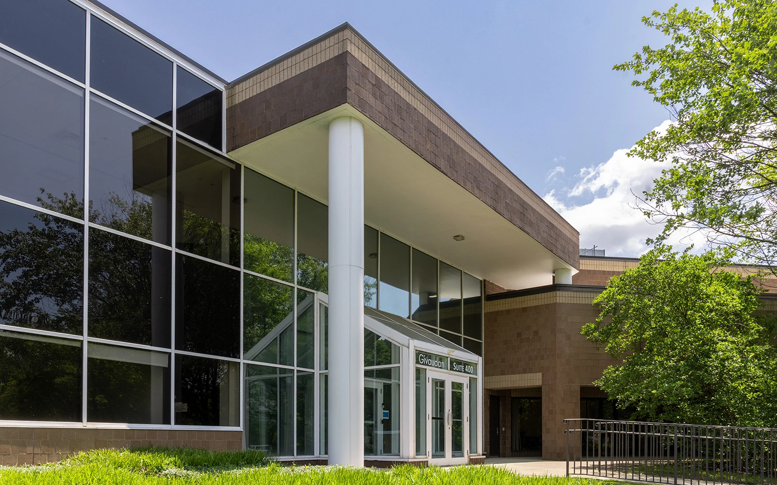 Modern office building entrance with glass facade columns and landscaped greenery under blue sky