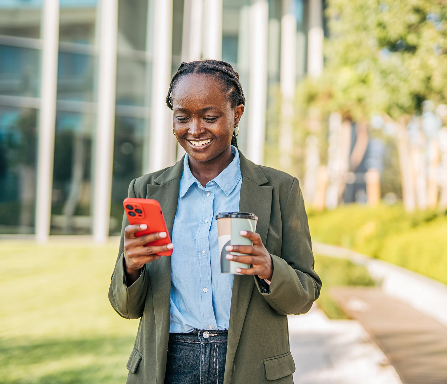 Professional woman smiling while checking smartphone and holding coffee outside modern office building